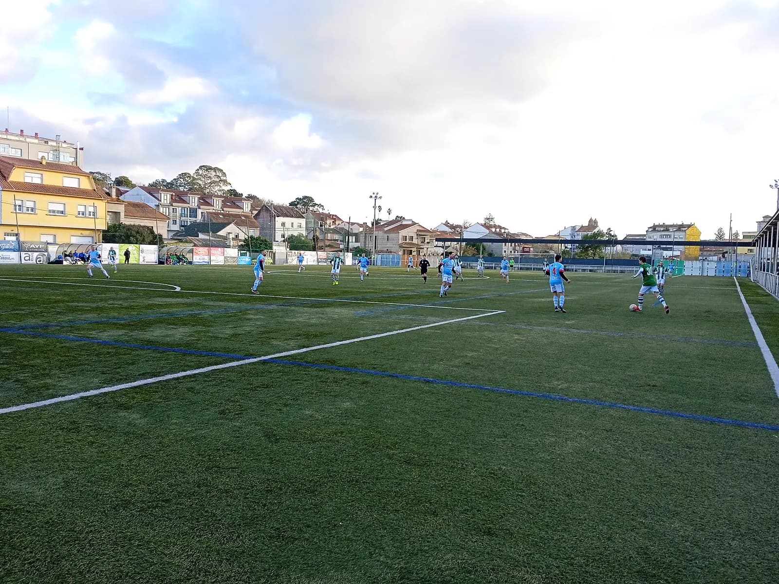 Partido en una cancha de pasto de pueblo, con casas detrás de la tribuna