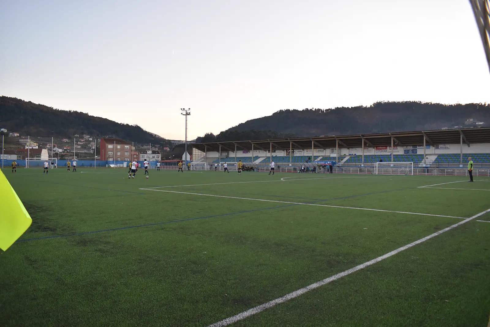 Cancha con una torre de reflectores y una tribuna techada, con los cerros detrás